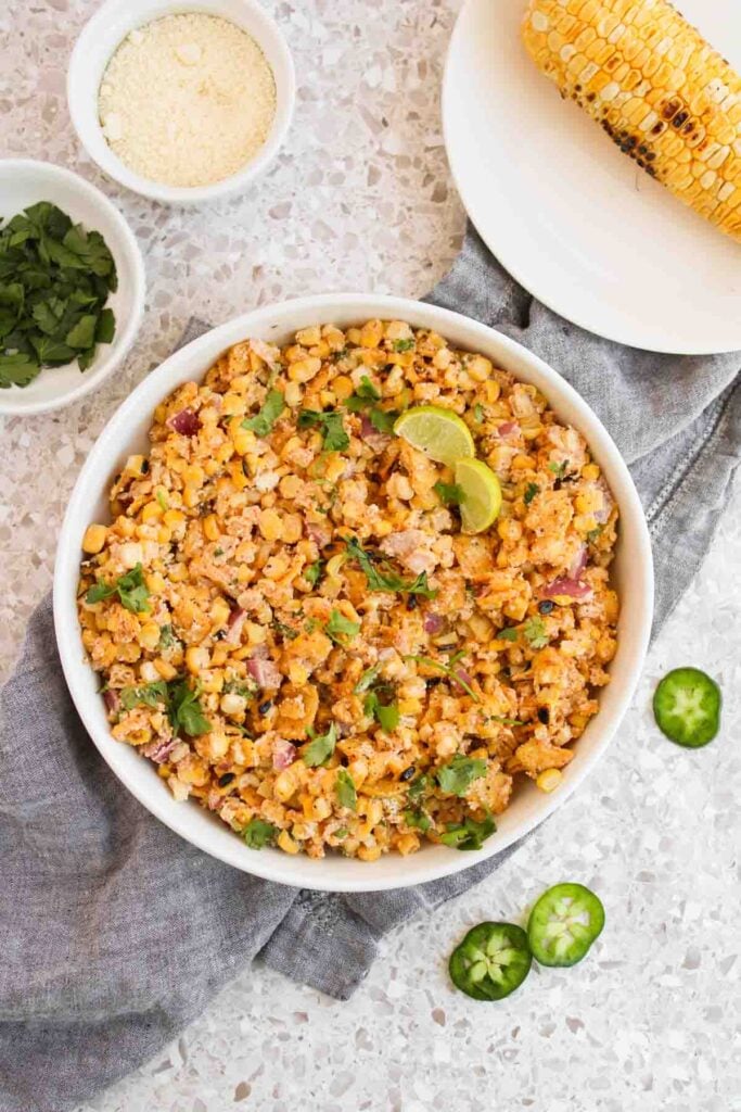 A bowl of corn salad with chopped cilantro and lime slices on top evokes the flavors of Mexican street corn. Nearby, a plate with grilled corn, a dish of grated cheese, and a bowl of chopped herbs complete this vibrant spread.