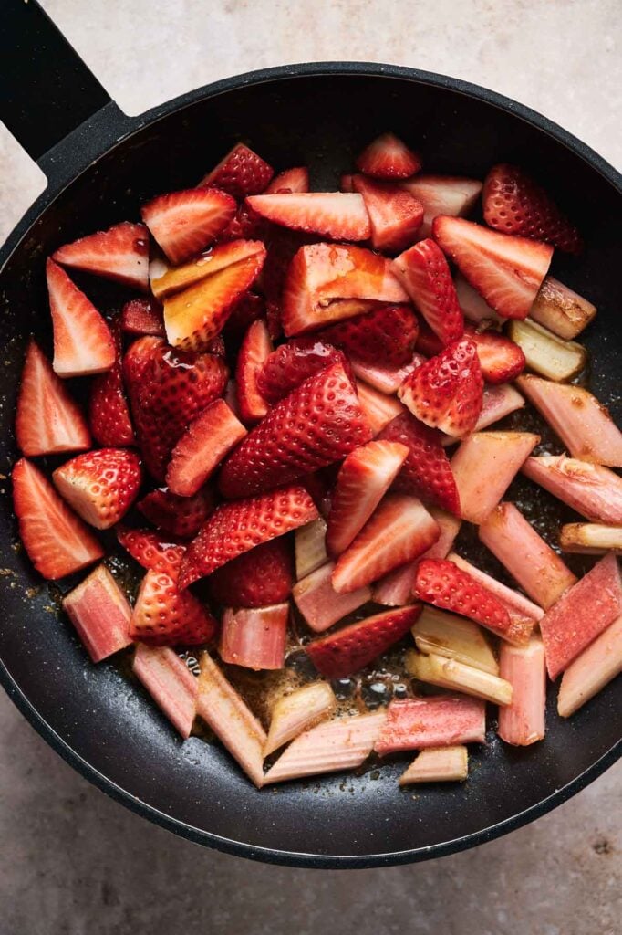Sliced strawberries and rhubarb simmer in a black pan, their juices mingling to create the perfect filling for a delightful rhubarb pie.