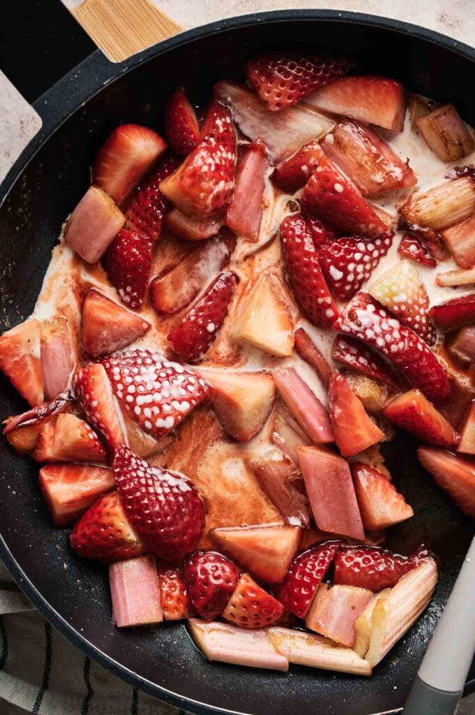 Chopped strawberries and rhubarb sizzle in a skillet, creating the beginnings of a delightful syrupy sauce reminiscent of a classic rhubarb pie.