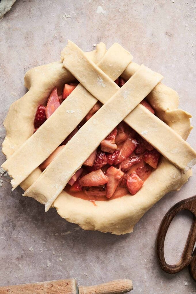 A rhubarb pie with a raw lattice crust, brimming with sliced fruit, rests on a floured surface. Nearby, scissors and wooden handles wait at the ready.