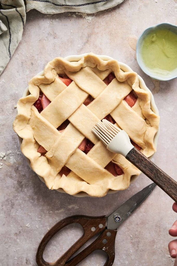 A person brushes egg wash on a lattice-topped rhubarb pie crust. Scissors and a small bowl sit nearby on the marble surface.