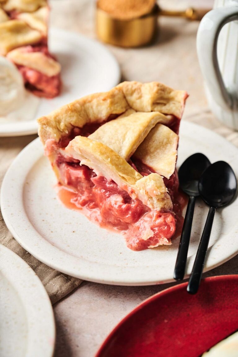 A slice of rhubarb pie with a golden crust and pink filling rests on a white plate. Two black spoons are placed beside it.