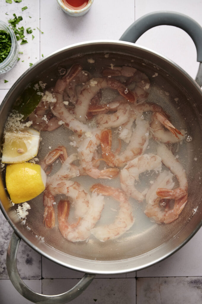Shrimp cooking in a pot with water, lemon wedges, garlic, and a bay leaf evokes the start of a delicious shrimp cocktail, viewed from above on a tiled surface.