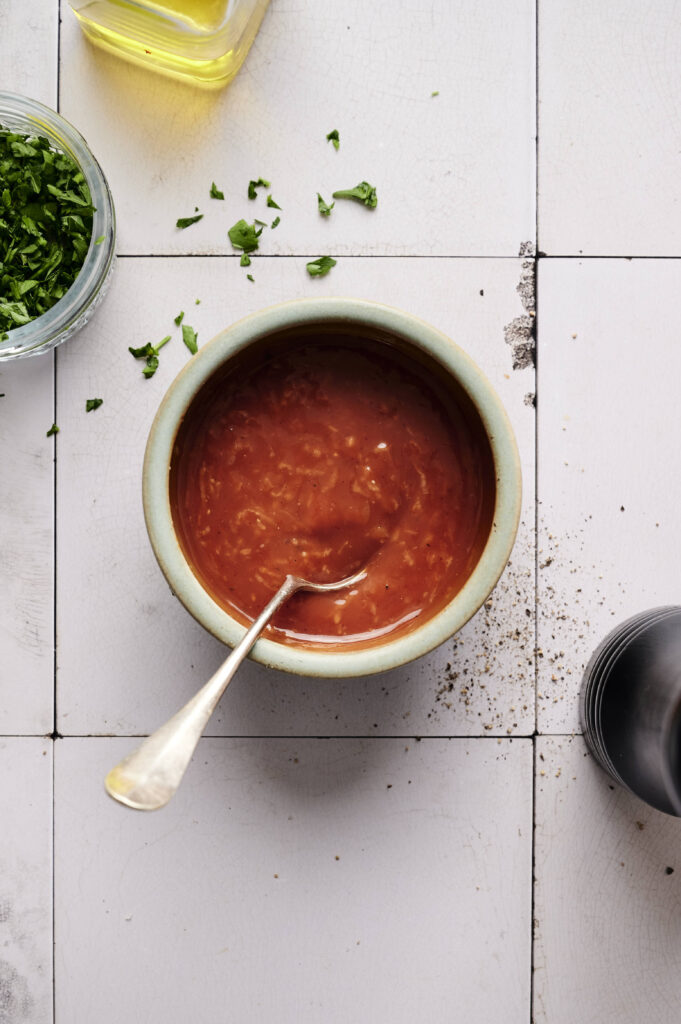 A bowl of red sauce, reminiscent of a tangy shrimp cocktail, sits with a spoon on a tiled surface. Scattered herbs and a glass container with chopped greens accompany it, while the edge of a bottle peeks in.
