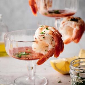 Three shrimp cocktails in glass cups, beautifully garnished with fresh herbs, sit elegantly alongside lemon wedges and a nearby jar of herbs on a pristine white surface.