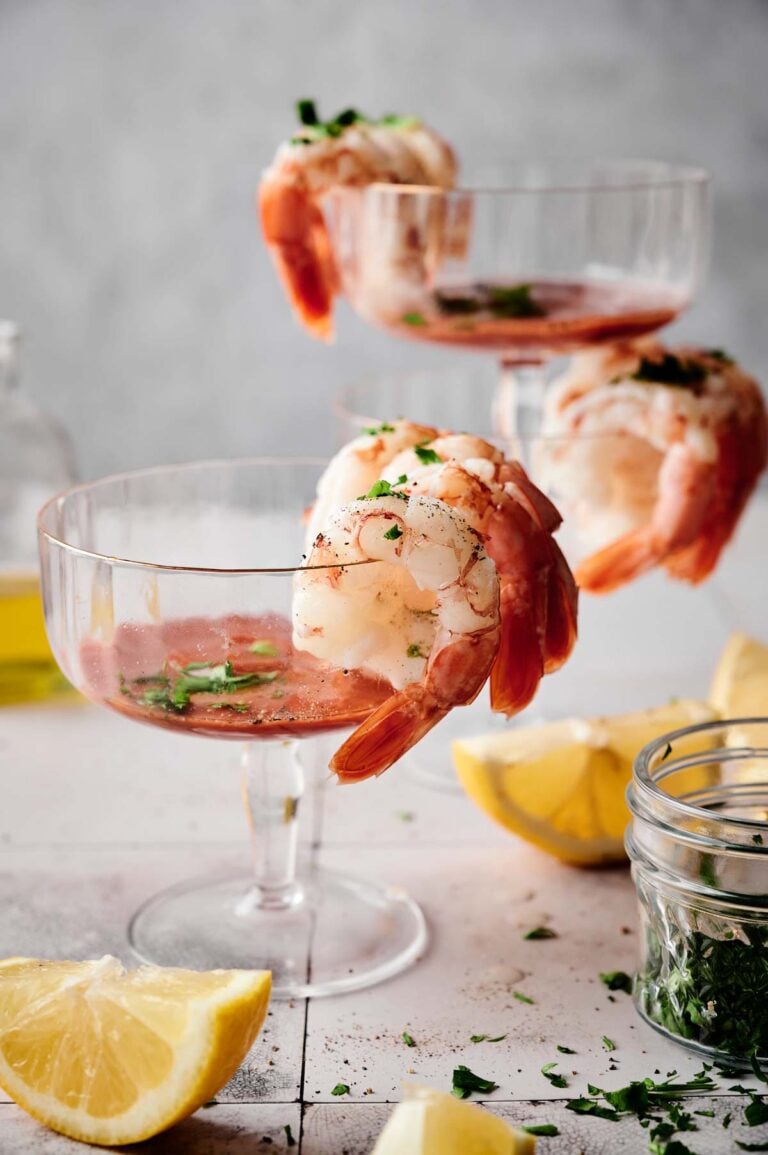 Three shrimp cocktails in glass cups, beautifully garnished with fresh herbs, sit elegantly alongside lemon wedges and a nearby jar of herbs on a pristine white surface.
