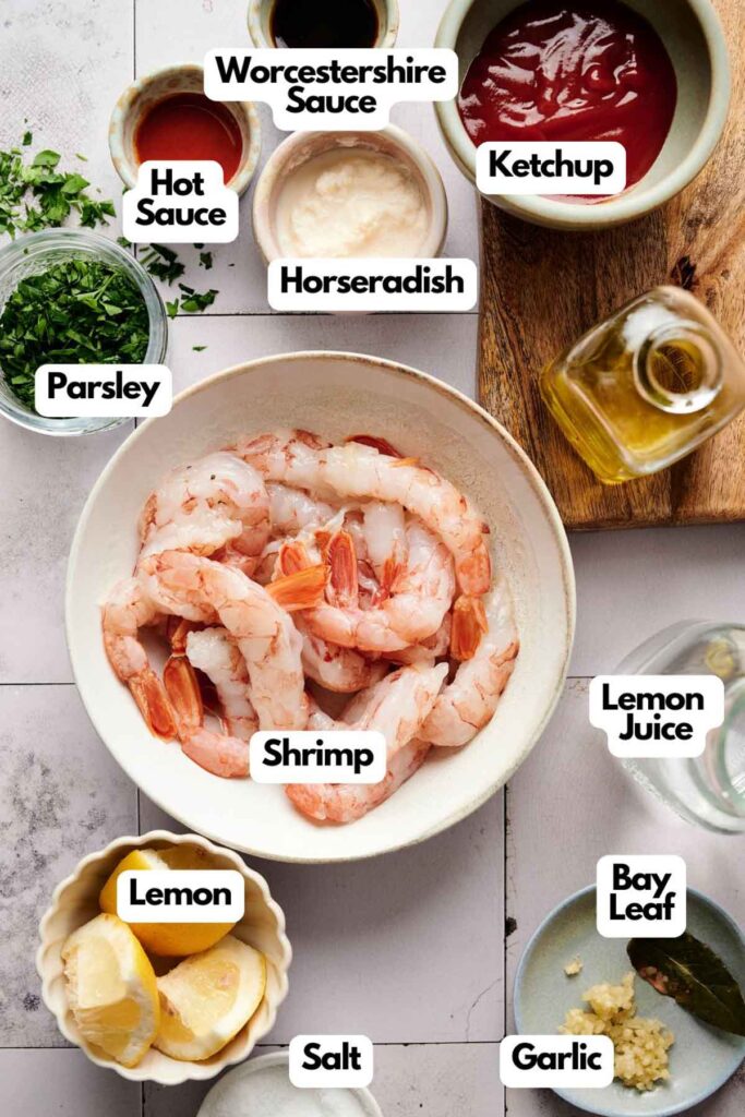 A shrimp cocktail awaits, with ingredients like shrimp, ketchup, Worcestershire sauce, hot sauce, and horseradish laid out. Parsley, lemon juice, salt, garlic, a bay leaf, and olive oil complete this seafood delight on the table.