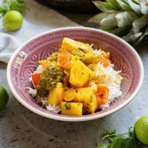A bowl of rice topped with a vibrant vegetable Thai coconut curry, featuring broccoli and pineapple chunks. Lime and cilantro garnish the table.