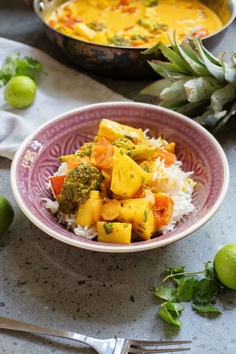 A bowl of rice topped with a vibrant vegetable Thai coconut curry, featuring broccoli and pineapple chunks. Lime and cilantro garnish the table.