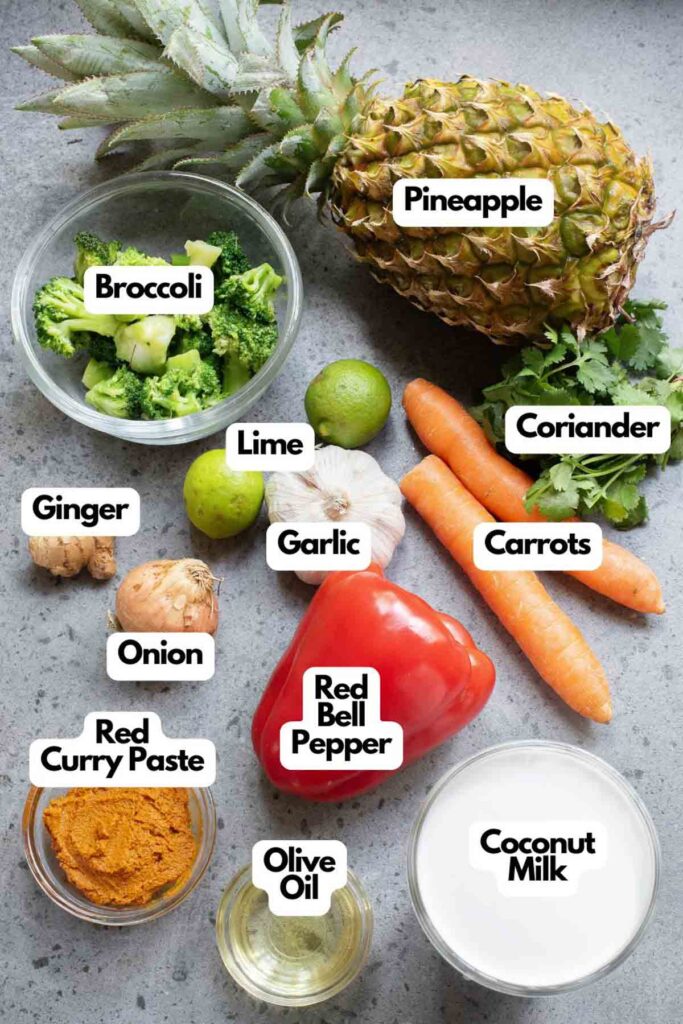 Top view of various labeled ingredients for Thai Coconut Curry, featuring pineapple, broccoli, lime, ginger, garlic, onion, red curry paste, olive oil, red bell pepper, coconut milk, carrots, and coriander.