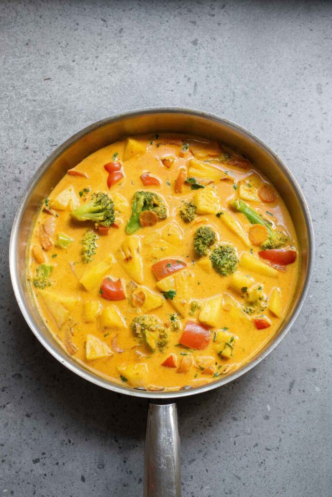 A pan filled with Thai Coconut Curry, featuring a vibrant mix of broccoli, red bell peppers, and diced potatoes, sits enticingly on a gray surface.