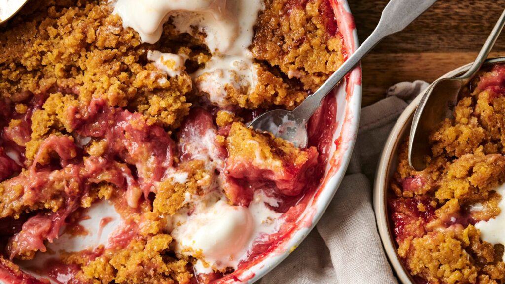 A dish of peach and raspberry crumble is topped with melting vanilla ice cream. A small portion has been scooped into a bowl on the side. Two spoons are visible.