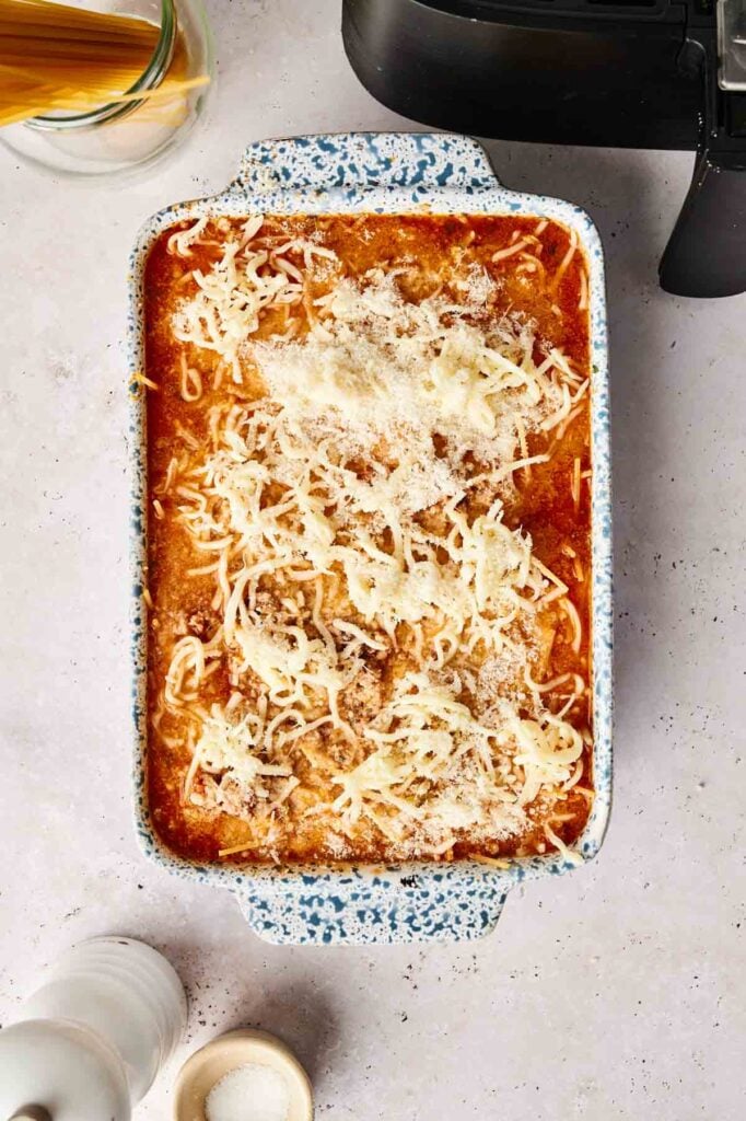 A rectangular dish brimming with creamy air fryer spaghetti casserole, crowned with shredded cheese and grated parmesan, rests invitingly on a light countertop beside a pepper grinder.