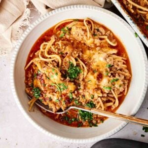 A bowl of air fryer spaghetti casserole with ground meat, melted cheese, and chopped parsley. A fork rests in the bowl, and a beige cloth napkin is beside it.