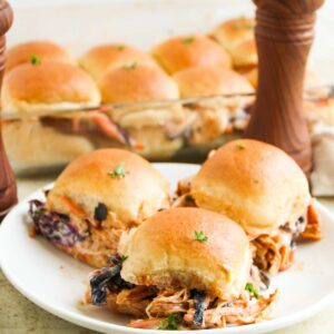 Three BBQ Chicken Sliders topped with slaw on a white plate, with a dish of extra sliders and two wooden pepper mills in the background.