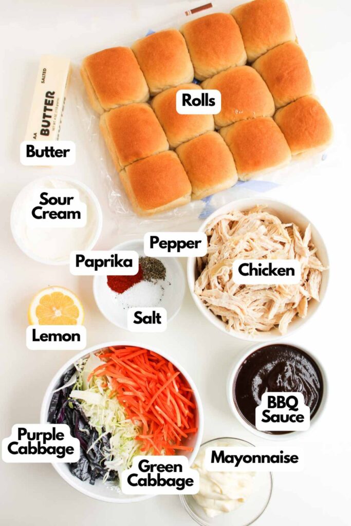 Ingredients for BBQ Chicken Sliders are arranged on a white surface, including rolls, shredded chicken, BBQ sauce, cabbage, carrots, seasonings, mayonnaise, sour cream, lemon, and butter.