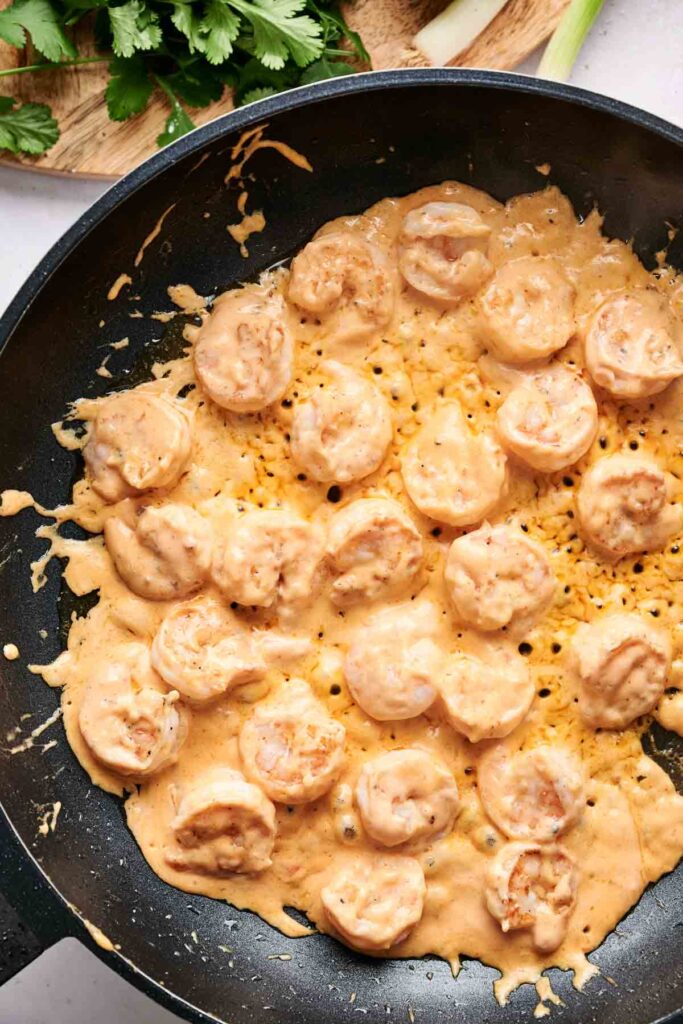 Shrimp cooked in a creamy, light orange sauce in a black skillet, with green herbs partially visible in the background.