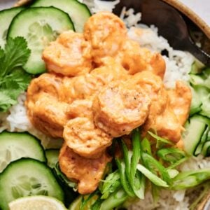 A bowl of rice topped with spicy shrimp, cucumber slices, and green onions, garnished with lime wedges and cilantro.