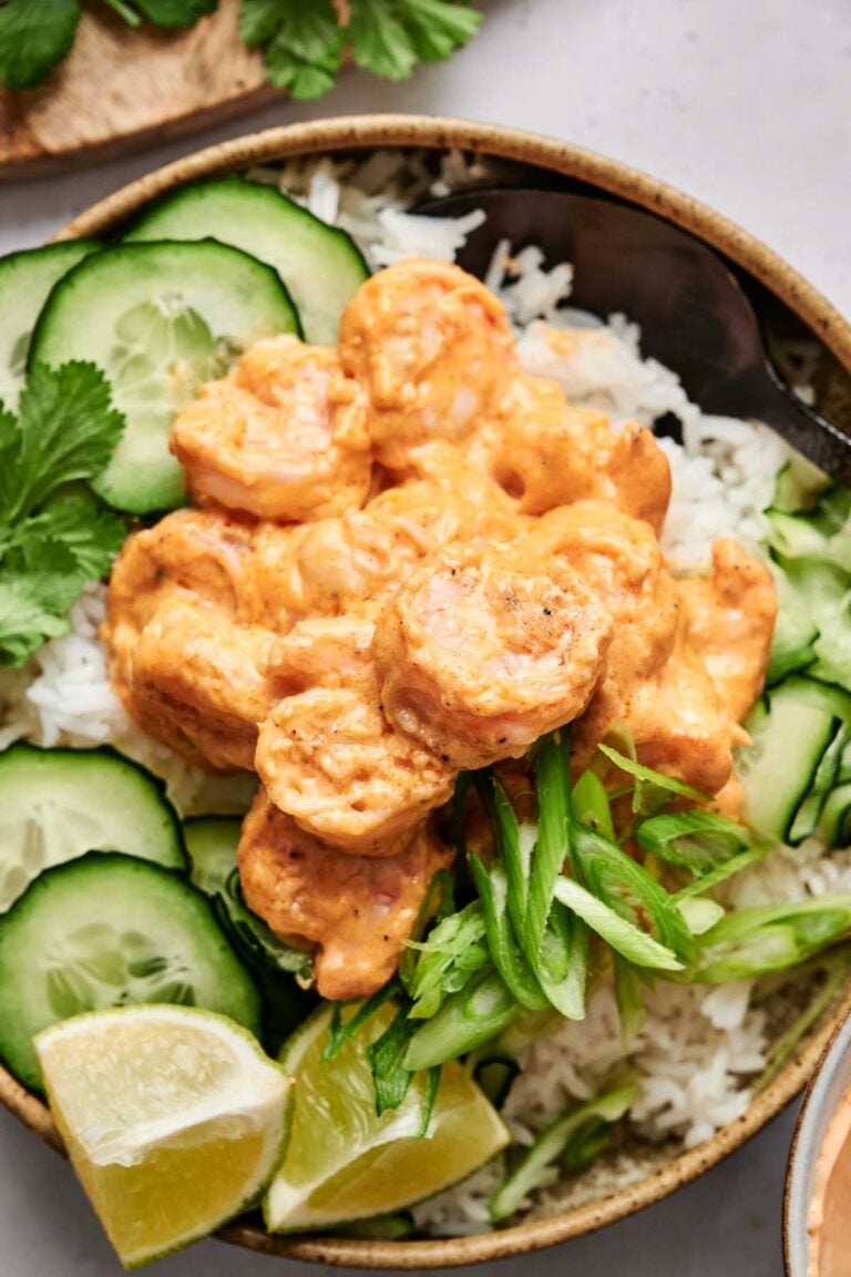 A bowl of rice topped with spicy shrimp, cucumber slices, and green onions, garnished with lime wedges and cilantro.