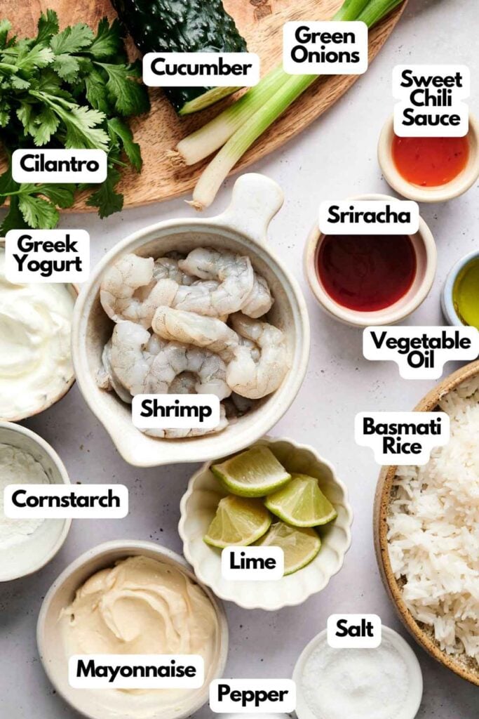 Ingredients for a recipe are arranged: shrimp, Greek yogurt, mayonnaise, cornstarch, sriracha, sweet chili sauce, vegetable oil, lime, cucumber, green onions, cilantro, salt, pepper, and basmati rice.