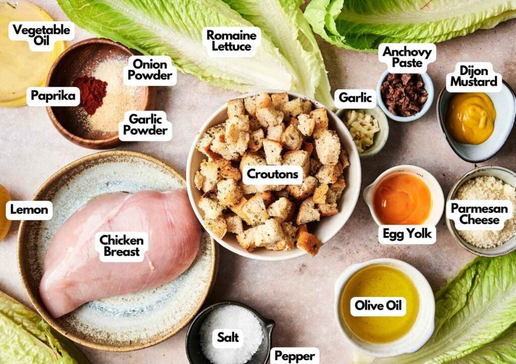 A delicious Chicken Caesar Salad spread with labeled ingredients on a table, featuring chicken breast, lettuce, croutons, seasonings, egg yolk, oils, parmesan cheese, and condiments.