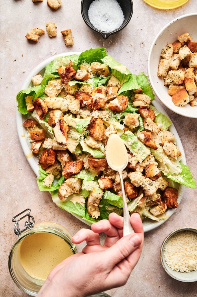 A hand drizzling dressing over a Chicken Caesar Salad with croutons and romaine lettuce on a platter. Surrounding the plate are bowls of croutons, grated cheese, and salt.