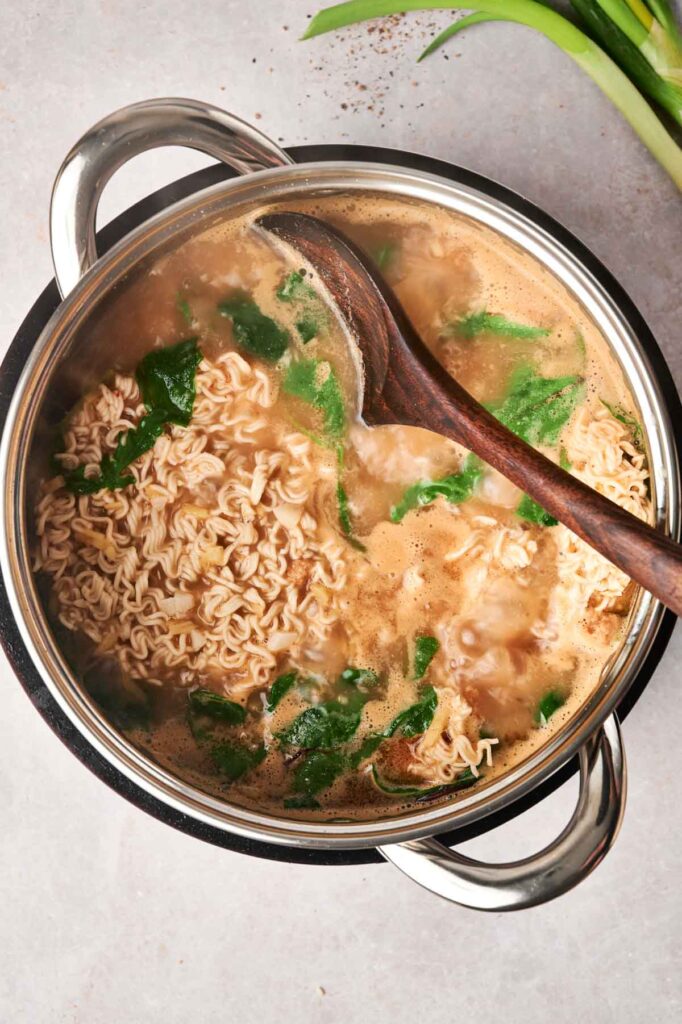 A pot of chicken ramen noodles boiling with green leafy vegetables, a wooden spoon resting on the rim, viewed from above.