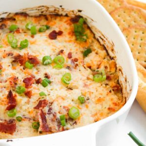 A white casserole dish holds the savory Crack Chicken Dip, crowned with melted cheese, crispy bacon bits, and a sprinkle of chopped green onions. On a wooden board nearby, crackers and a bowl of fresh green onions await to be paired with this irresistible delight.