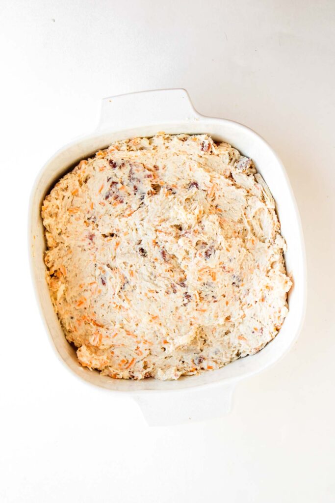 A square white dish filled with Crack Chicken Dip, showcasing a delightful mixture of shredded cheese and visible chunks, all set against a plain white background.
