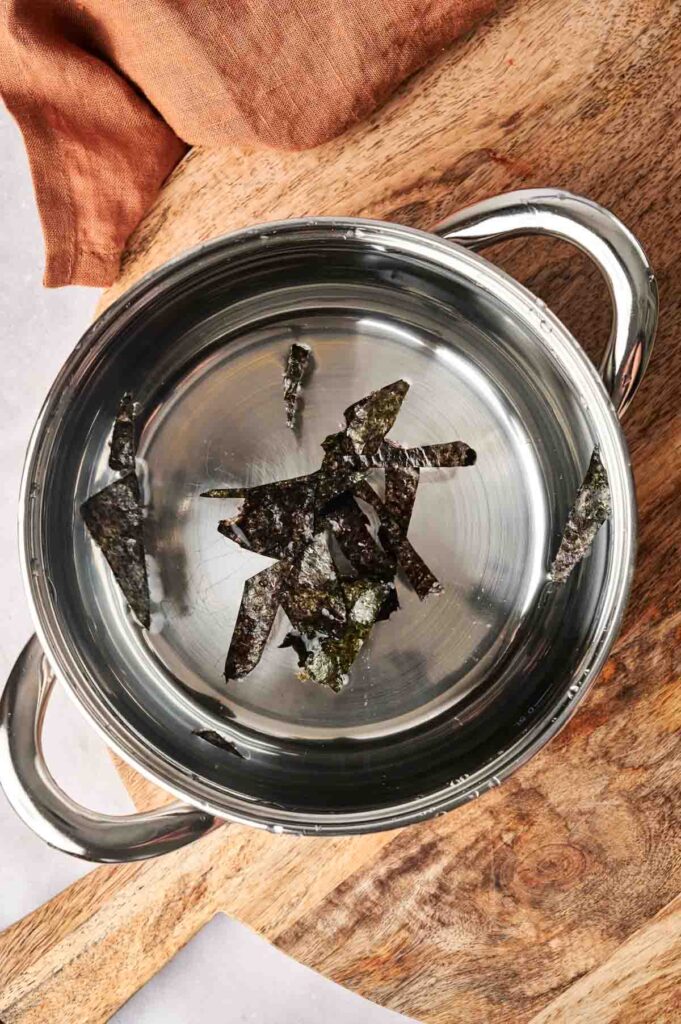 A silver pot filled with water and pieces of seaweed sits on a wooden surface, capturing the essence of dashi, while a brown cloth is draped elegantly beside it.
