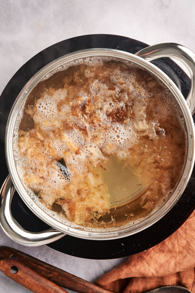 A pot of simmering dashi broth with visible herbs and spices on a stovetop, covered in frothy bubbles.