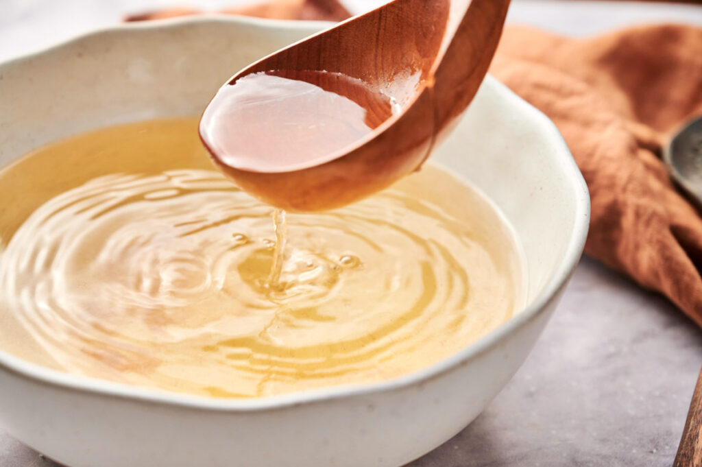 A wooden ladle pours rich dashi broth into a white bowl on a table.