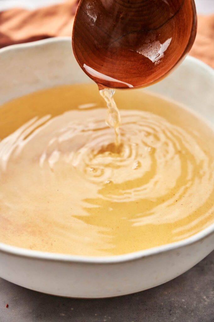 A wooden ladle gently pours dashi broth into a white bowl, creating ripples on the surface.