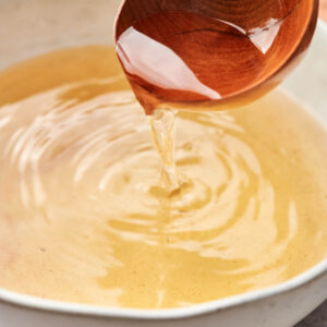 A wooden ladle pours clear dashi broth into a white bowl, creating gentle ripples on the surface.