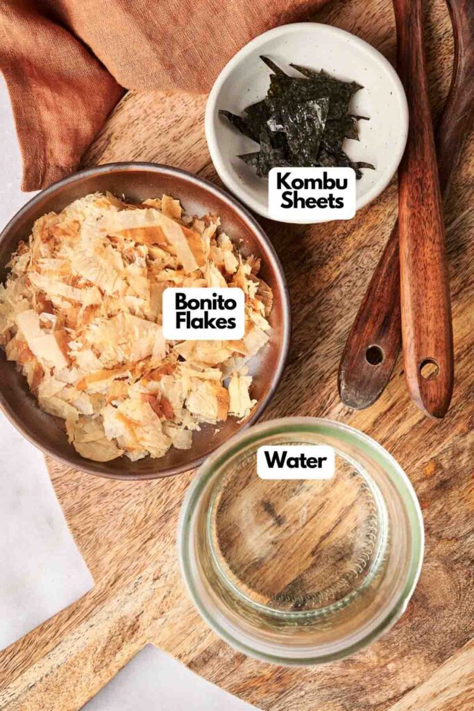 A wooden board holds a bowl of bonito flakes and a plate of kombu sheets, essential for crafting rich dashi. Nearby, a glass of water awaits its role in the process, with wooden utensils ready in the background.