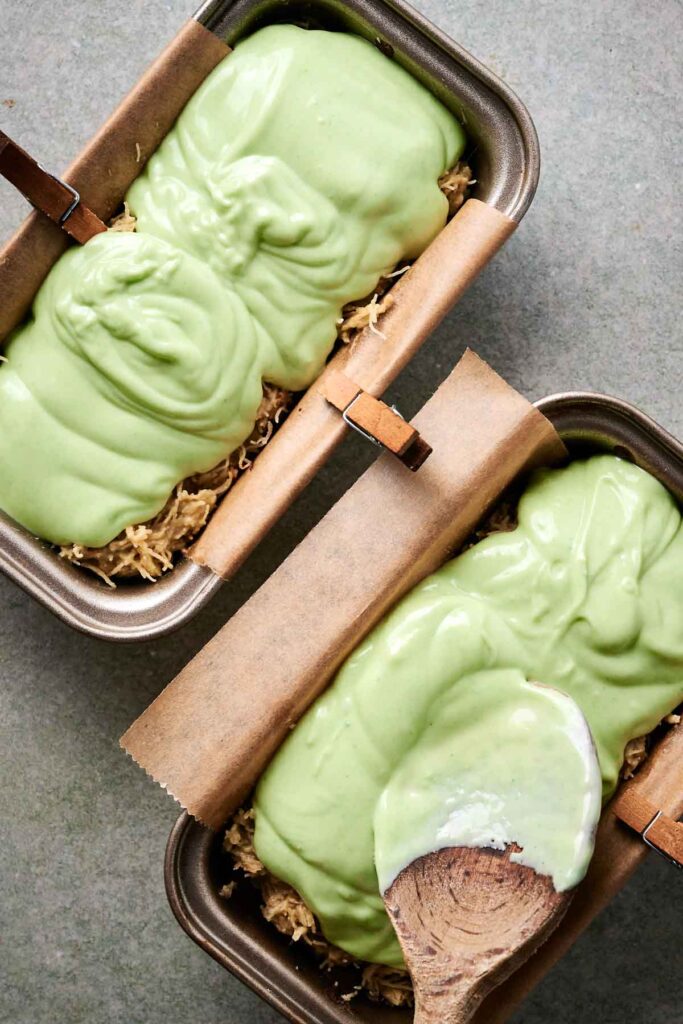 Two baking pans lined with parchment paper are filled with a mixture of ingredients. A creamy green topping, reminiscent of exotic Dubai chocolate, has been spread over each pan, with a wooden spoon resting on one.