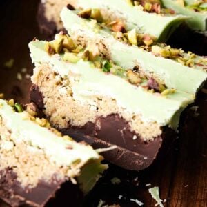 Sliced dessert bars featuring layers of luxurious Dubai chocolate, a crumbly filling, and a green topping sprinkled with chopped pistachios rest elegantly on a wooden surface.