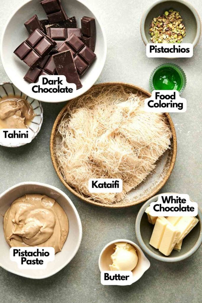 Ingredients arranged in bowls include dark chocolate, Dubai Chocolate, white chocolate, pistachios, tahini, pistachio paste, kataifi, butter, and food coloring on a gray surface.