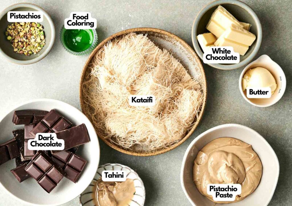 A top view of labeled ingredients in bowls for a dessert: pistachios, Dubai Chocolate, food coloring, kataifi, white chocolate, butter, dark chocolate, tahini, and pistachio paste.