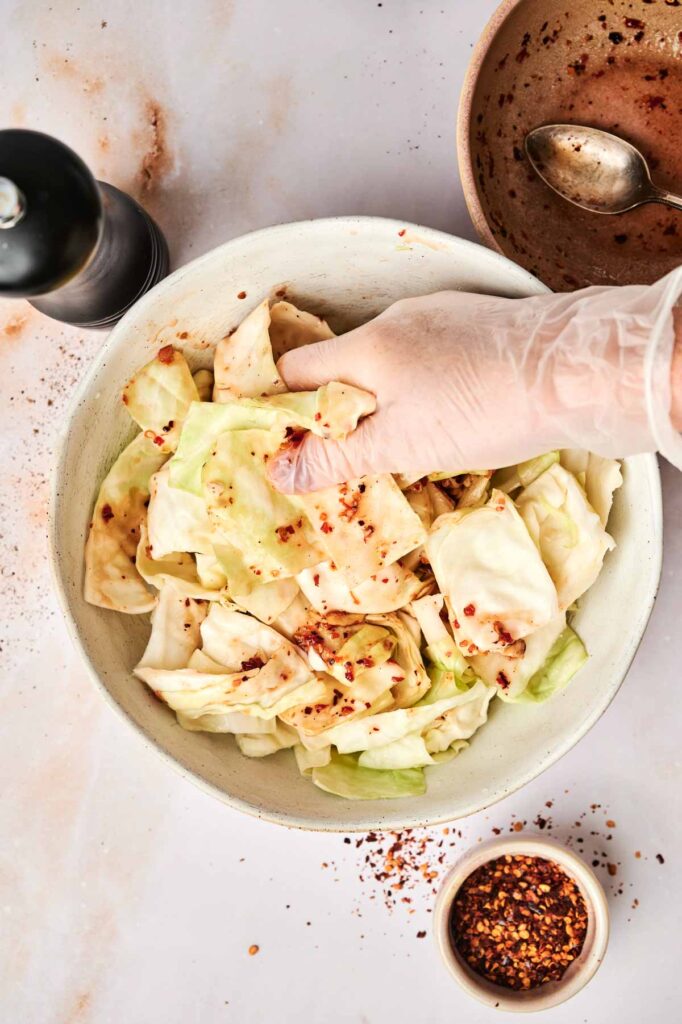 A gloved hand mixes chopped cabbage and red pepper flakes in a bowl, hinting at the start of homemade kimchi, with a pepper grinder and small bowl of red pepper flakes nearby.