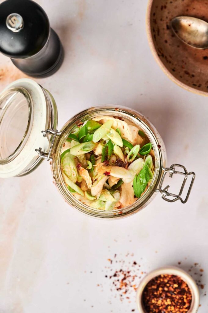 An open glass jar filled with sliced green onions, chicken pieces, and tangy kimchi sits on a light surface, surrounded by a pepper grinder, an empty bowl, and a small dish of red pepper flakes.