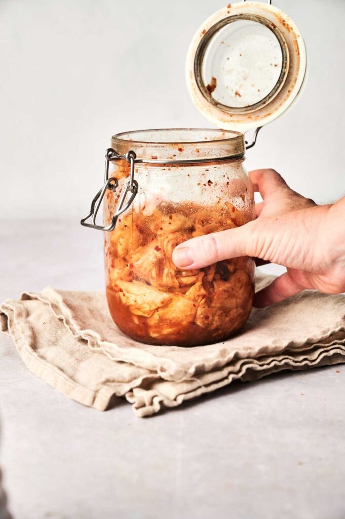 A hand opens a glass jar filled with vibrant kimchi, set on folded beige cloth napkins against a light background.