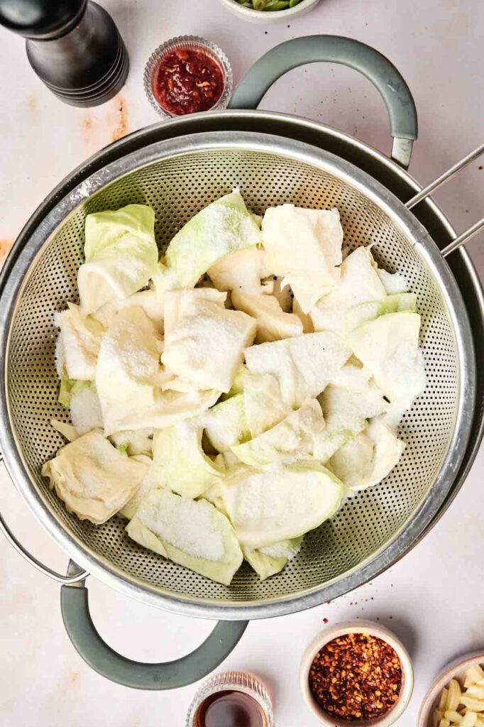 Chopped cabbage pieces sprinkled with salt in a colander, surrounded by small bowls of kimchi red pepper flakes, sauce, and other condiments.