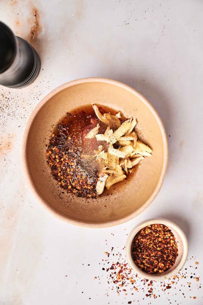A tan bowl containing shredded ginger, chili flakes, kimchi, sauce, and seasonings sits on a light surface next to a small dish of chili flakes and a pepper grinder.