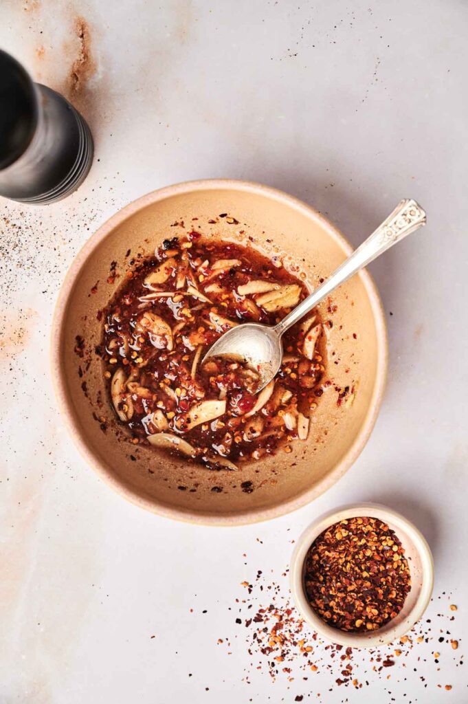 A beige bowl with chili oil, garlic slices, and kimchi sits on a white surface, accompanied by a small bowl of red pepper flakes and a pepper grinder.