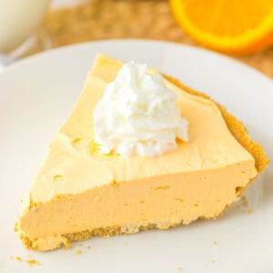 A slice of orange cream pie, reminiscent of a refreshing Kool Aid Pie, sits elegantly on a white plate. It's topped with whipped cream, with an orange slice and a glass of milk gently resting in the background.
