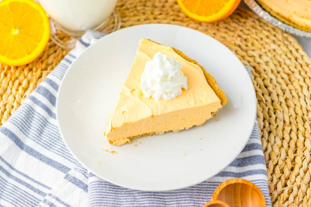A slice of Kool Aid Pie with whipped cream on top, served on a white plate alongside a striped napkin, fresh orange slices, and a glass of milk.