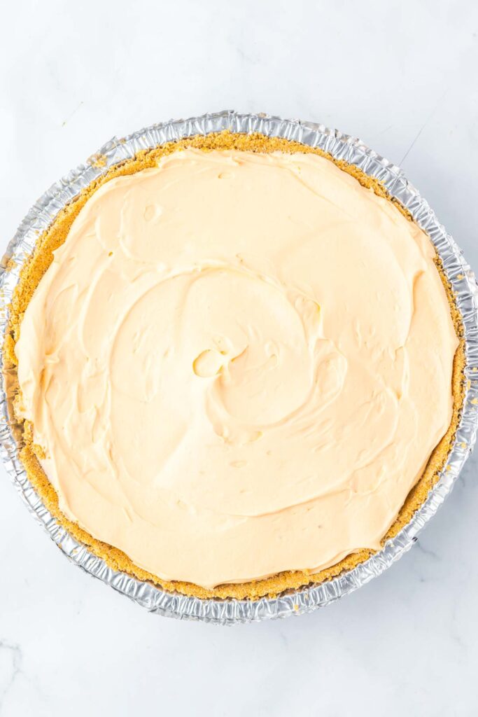 A round cheesecake reminiscent of a Kool Aid Pie sits in an aluminum pie pan, its smooth, light cream-colored top beautifully contrasting the white marble surface.