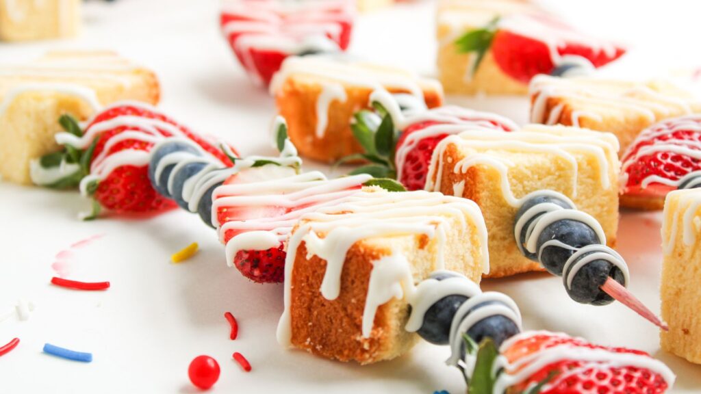 Skewers with pieces of pound cake, whole strawberries, and blueberries, drizzled with white icing, are arranged on a white surface with colorful sprinkles scattered around.