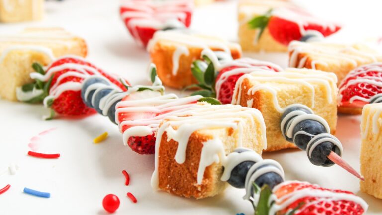 Skewers with pieces of pound cake, whole strawberries, and blueberries, drizzled with white icing, are arranged on a white surface with colorful sprinkles scattered around.